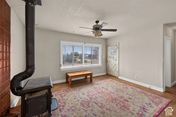 Hardwood flooring, wood-burning stove with copper-toned wall paneling, white-framed windows, and ceiling fan with integrated lighting