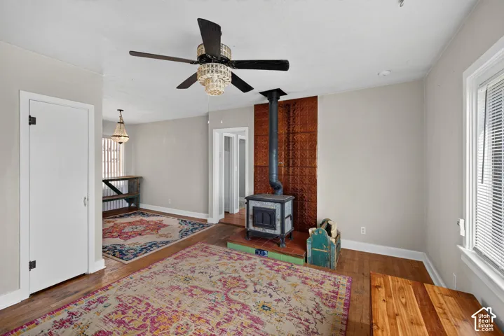 Wood-finish flooring, wood-burning stove with copper-toned backsplash, ceiling fan with chandelier lighting, and a white framed window with blinds