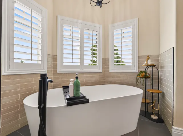 Freestanding soaking tub with matte black fixtures, multiple windows with plantation shutters, and floor-to-ceiling tile in a neutral tone