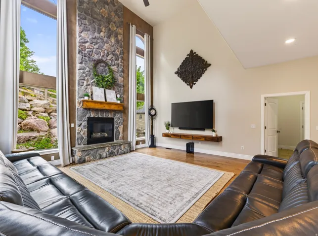 Vaulted ceiling, stone fireplace with wood mantel, large floor-to-ceiling windows, wood-finish flooring, and recessed lighting