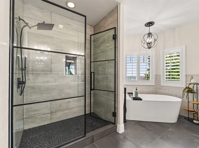 Spa-inspired primary bathroom featuring a frameless glass-enclosed walk-in shower with linear rainfall showerhead, dark tile flooring, and a freestanding soaking tub