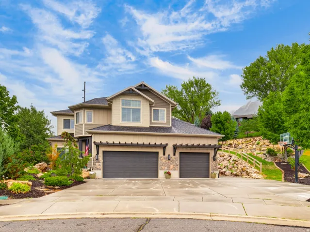 Two-car garage with dark composite doors and stone-accented facade, circular driveway with concrete surface, and mature landscaping including established trees and manicured garden beds throughout the property
