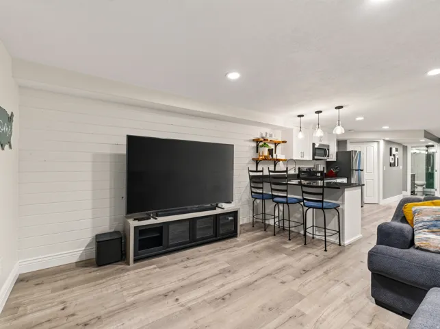 Wood-finish flooring throughout, white shiplap accent wall, recessed lighting, kitchen island with dark countertop, stainless steel appliances, and white cabinetry
