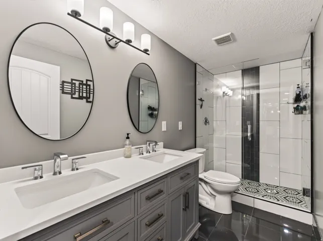 Spa-inspired primary bathroom featuring dual vanities with soft-close cabinetry and quartz countertops, frameless glass-enclosed shower with subway tile surround and mosaic floor accent, polished black tile flooring, and dual round mirrors with integrated