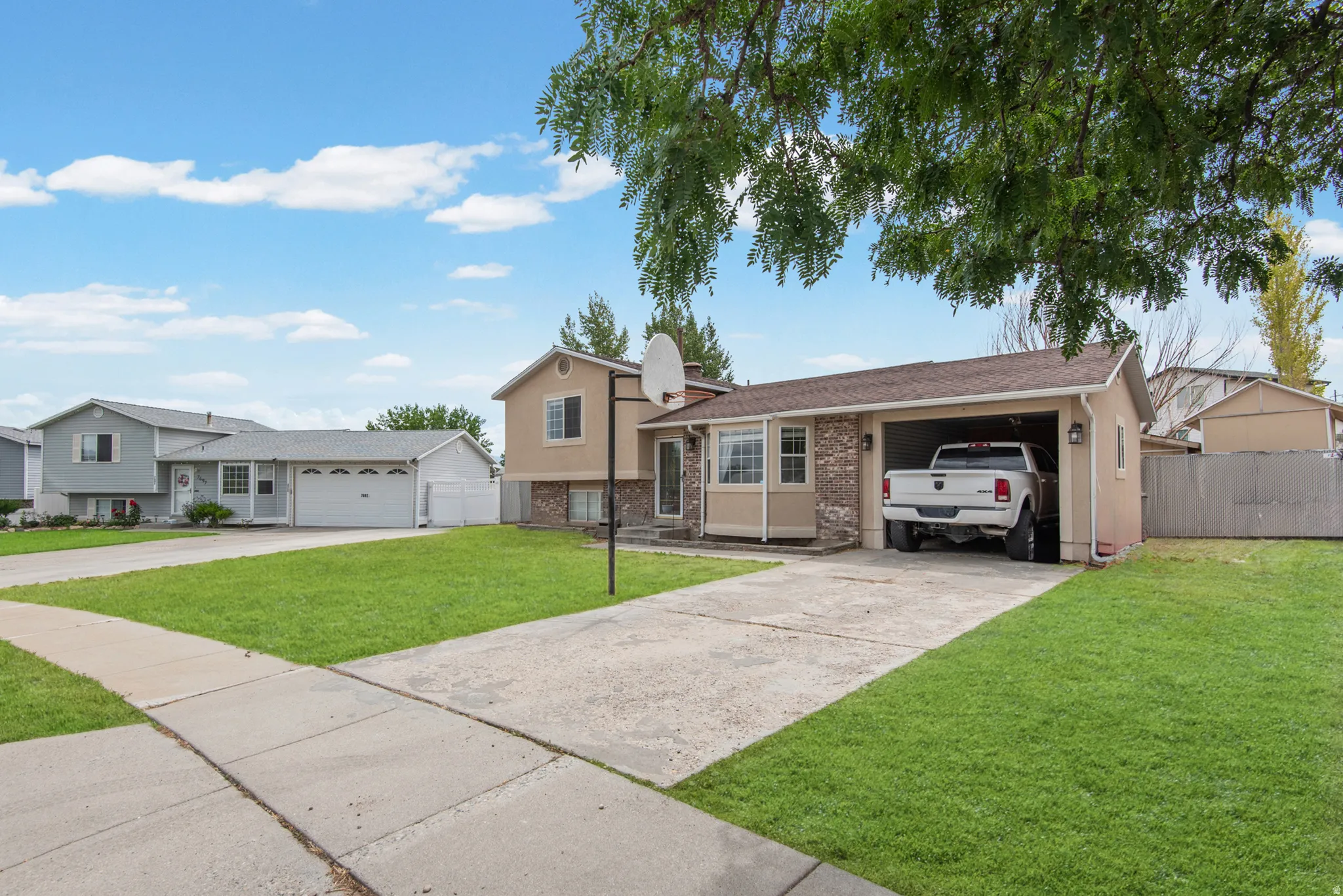 Welcoming single-story home featuring a spacious driveway with carport parking, manicured front lawn, mature landscaping with established trees, concrete walkway, and neighboring properties characteristic of a residential community setting