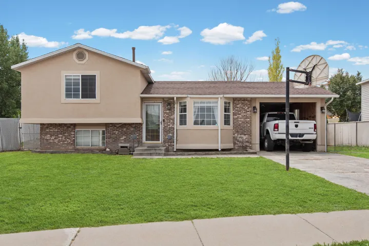 Welcoming split-level home featuring a brick and stucco exterior, attached carport with driveway, expansive manicured lawn, mature landscaping with poplar trees, and a basketball hoop for recreational use