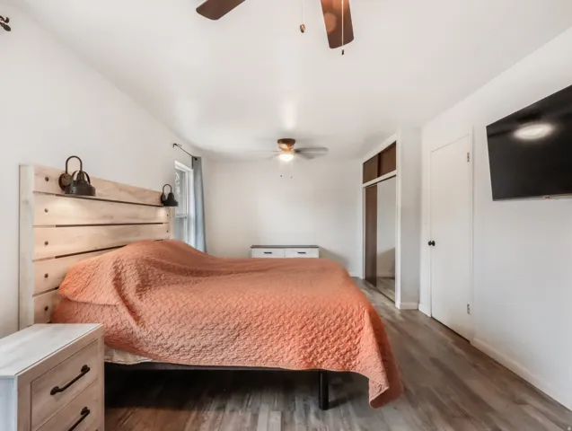 Wood-finish flooring, ceiling fan, white paneled door, mirrored closet doors, and window with curtain rod