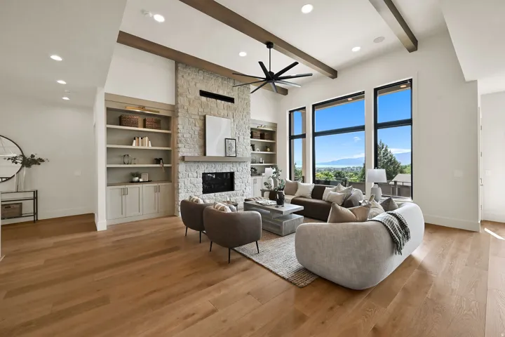 Vaulted ceiling with exposed wood beams, stone-clad fireplace, built-in shelving with lower cabinetry, wood-finish flooring, and expansive windows