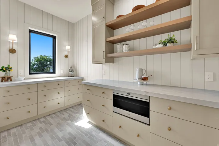 Light-toned cabinetry with brass hardware, wood-finish open shelving, integrated microwave, window with black trim, and plank wall paneling