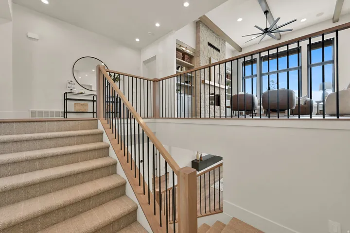Carpeted staircase with light wood handrail and black metal balusters, recessed lighting, and a prominent open living area featuring large windows
