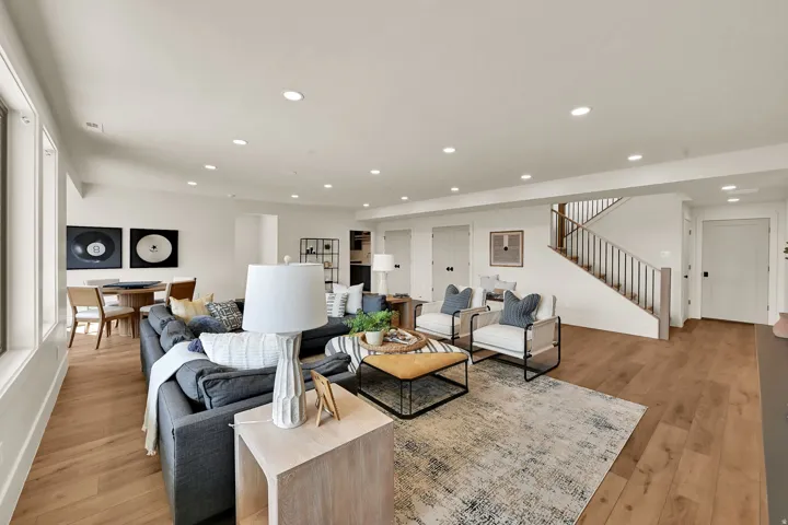 Spacious open-concept living area featuring wood-finish flooring, recessed lighting, multiple windows, and a prominent staircase with wood railings
