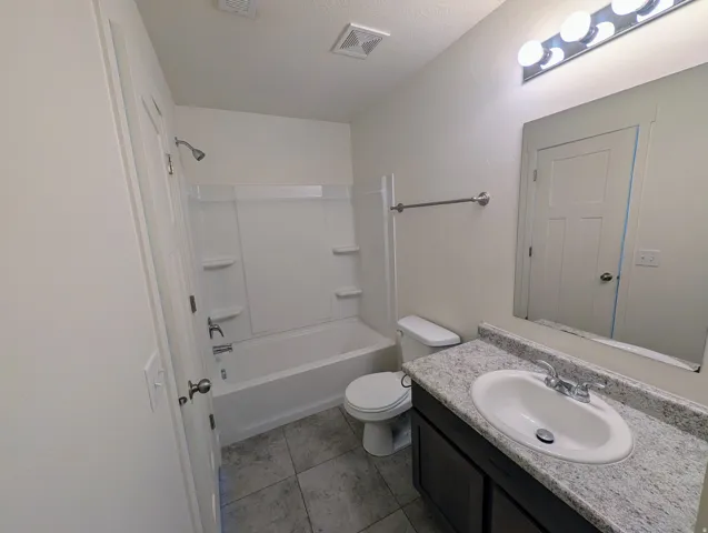 Bathroom featuring a white bathtub and shower combination, dark wood-finish vanity with an integrated sink, textured light-toned countertop, and gray tile flooring