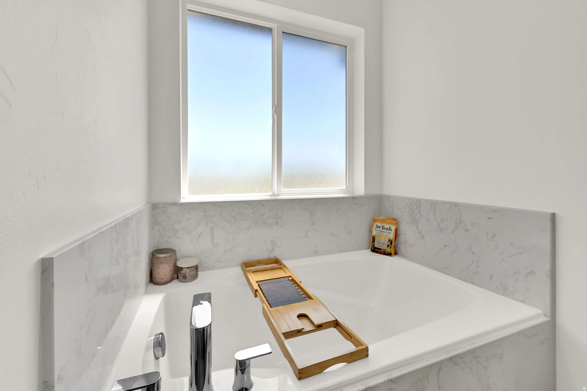 Built-in soaking tub with chrome fixtures, surrounded by white-veined surfacing, and a frosted window