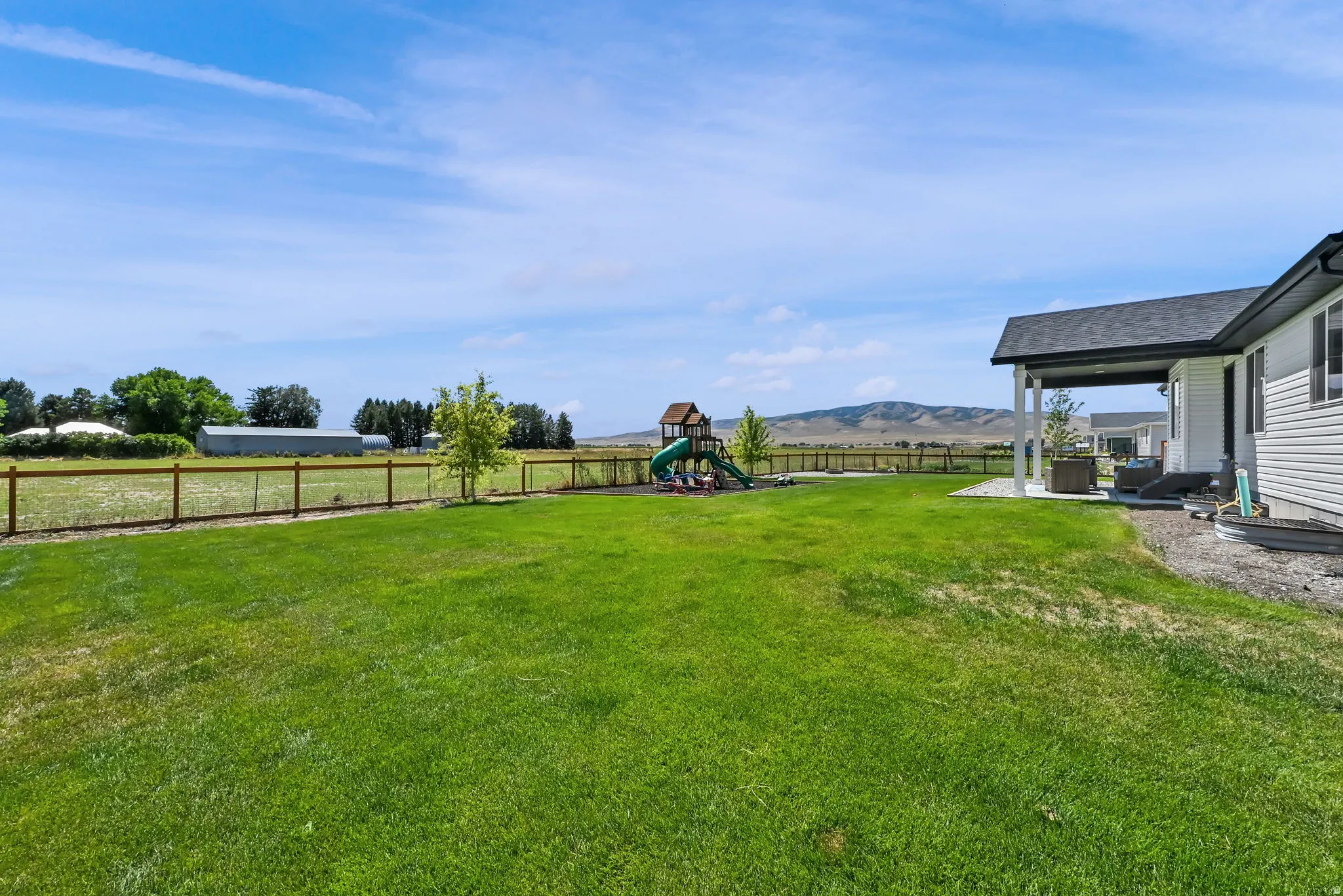 Expansive grassy yard with a covered patio, wood and wire fence, and distant mountain backdrop
