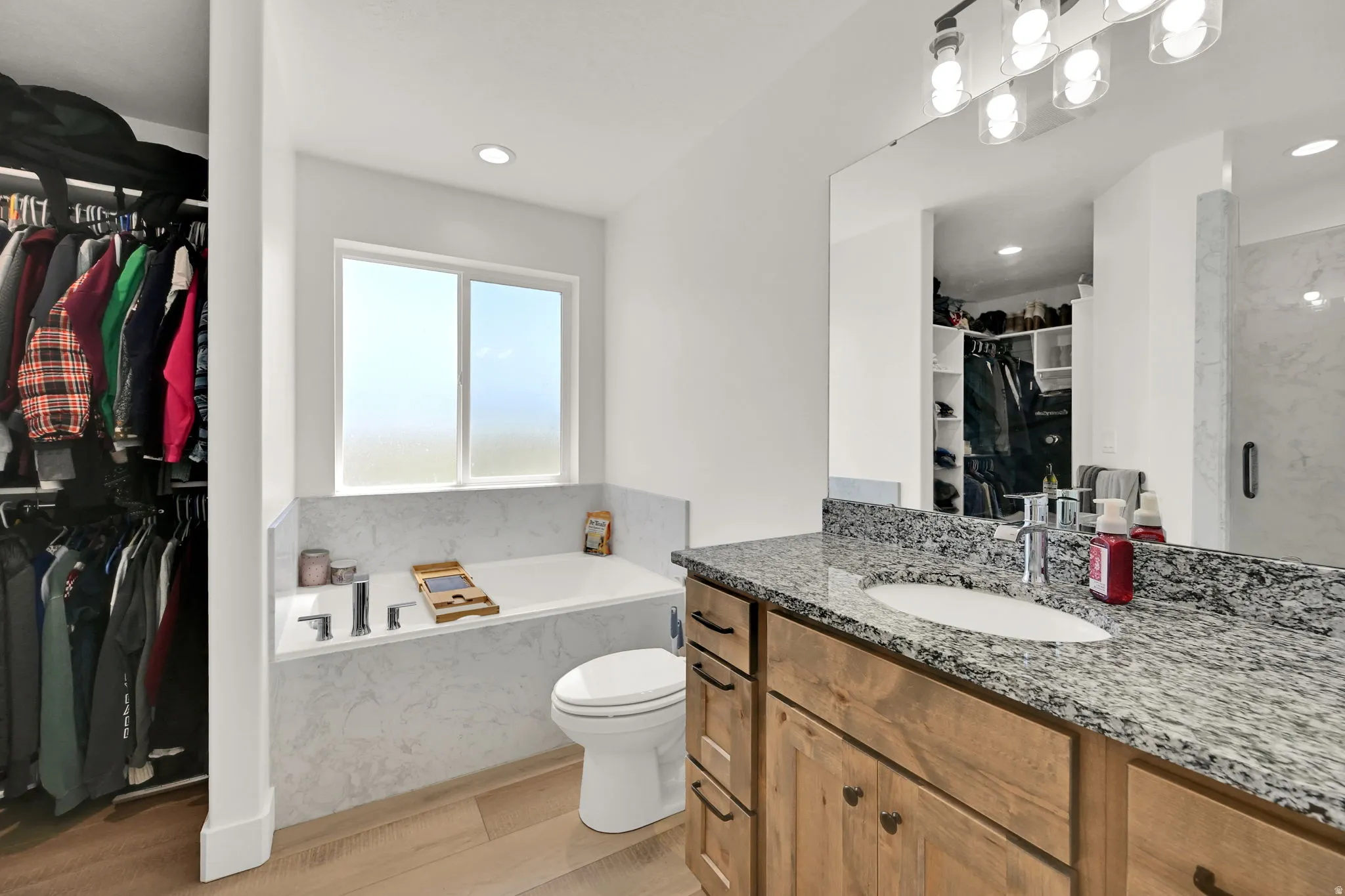 Built-in soaking tub with a tiled surround, recessed lighting, wood-finish flooring, vanity with a granite countertop, and integrated closet shelving
