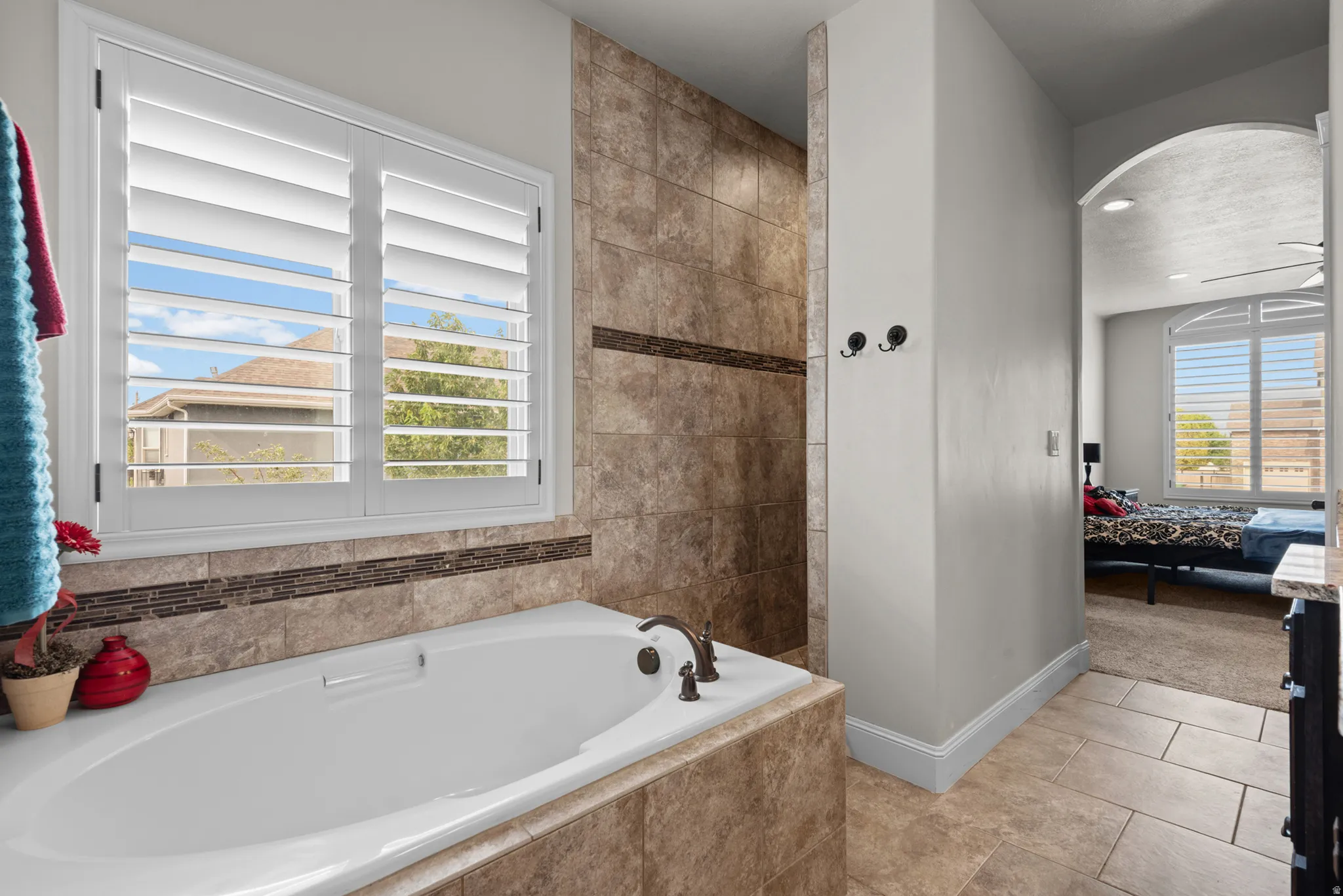 Soaking tub with bronze fixtures, ceramic tile surround with decorative accent, window with plantation shutters, tiled shower enclosure, and arched doorway