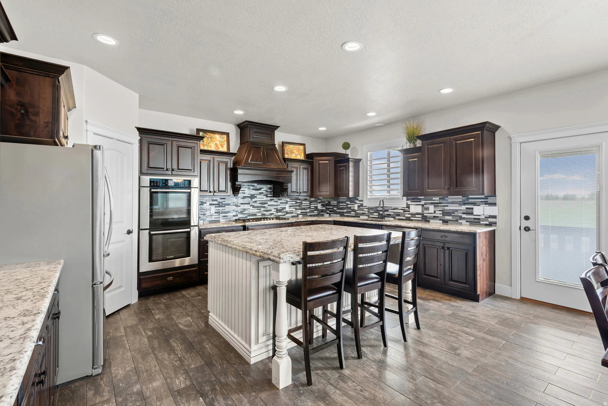 Chef's kitchen featuring custom dark cabinetry with glass-front upper displays, stainless steel double wall ovens, a professional-style range hood, quartz countertops, and a large center island with seating