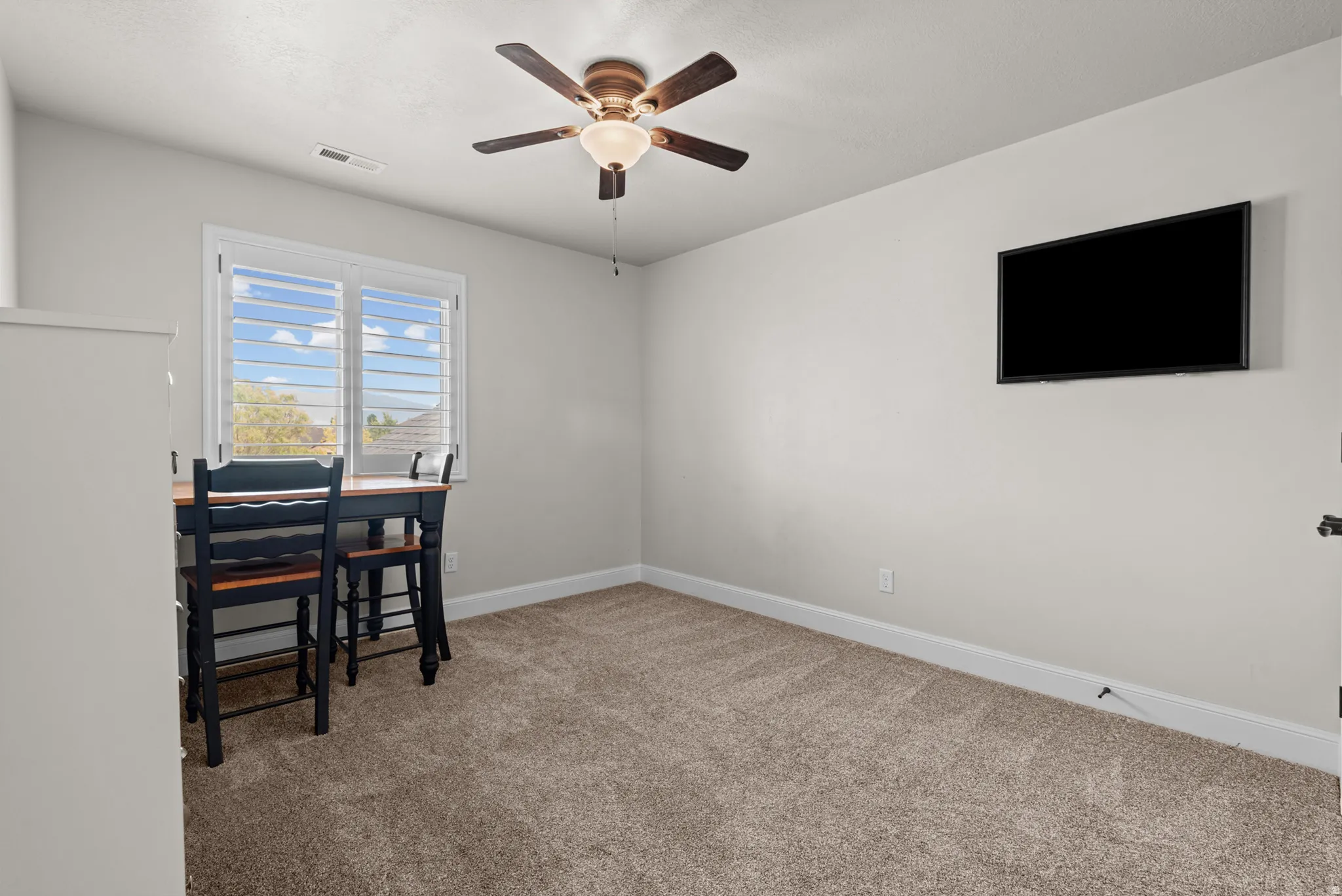 Carpeted room with a ceiling fan, window with white louvered shutters, and a wall-mounted display