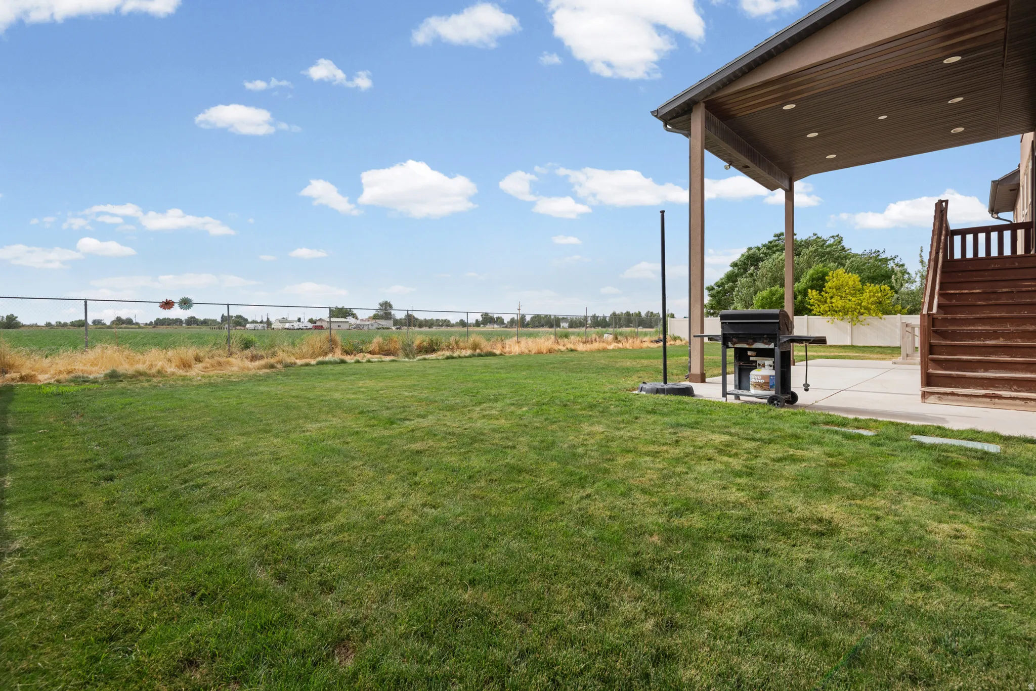 Expansive grass lawn with a covered concrete patio, wood deck, and chain link fencing