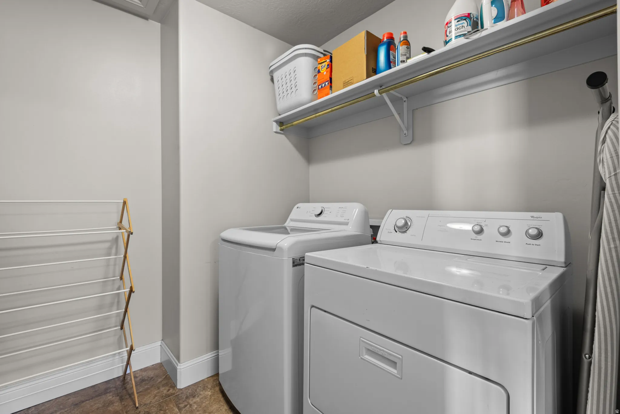 Dedicated laundry room featuring side-by-side washer and dryer with white finishes, stainless steel shelving with brass hardware for storage, wood-finish flooring, and a wall-mounted drying rack
