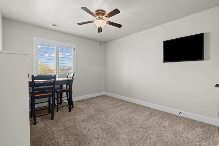 Carpeted room with a ceiling fan, window with white louvered shutters, and a wall-mounted display