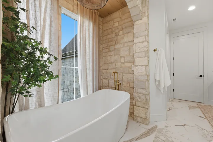 Freestanding soaking tub with floor-mounted faucet, stone accent wall, wood-paneled ceiling, and large window with privacy drapes