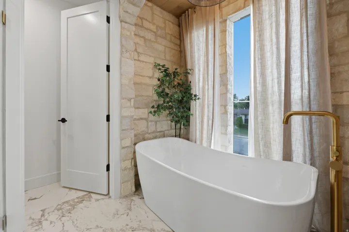 Freestanding soaking tub with a floor-mounted golden faucet, stone accent wall, and large window with drapes