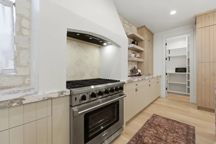 Stainless steel range, decorative hood, patterned tile backsplash, light wood-finish flooring, and a walk-in pantry with built-in shelving