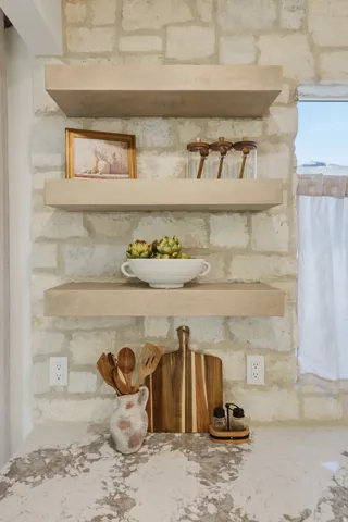 Stone wall with three light-toned floating shelves, a patterned countertop, and a window with a sheer curtain