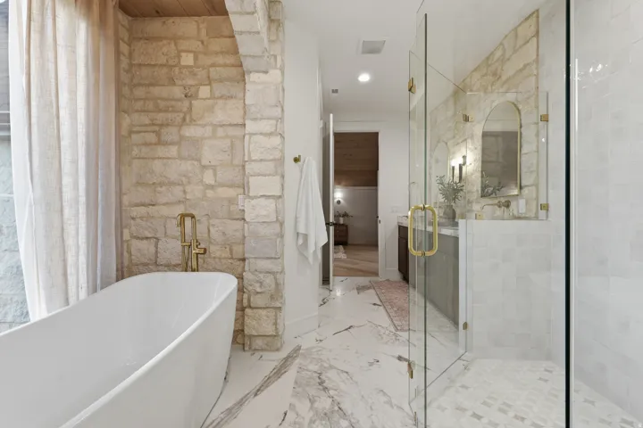 Freestanding soaking tub with a floor-mounted brass faucet, stone accent walls, a glass-enclosed shower, a double vanity with an arched mirror, and marble-finish flooring