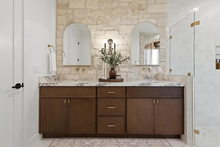 Dual sink vanity with stone countertop, wood-finish cabinetry, and stone-clad accent wall
