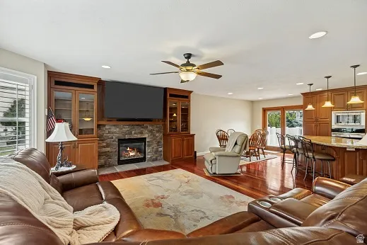 Built-in cabinetry with glass-front display, stone-clad fireplace, Brazilian Cherry wood-finish flooring, recessed lighting, and a ceiling fan