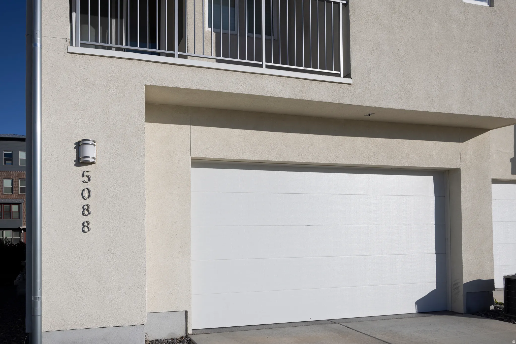 Smooth stucco exterior with a white garage door, modern exterior lighting, and an upper-level balcony with metal railing