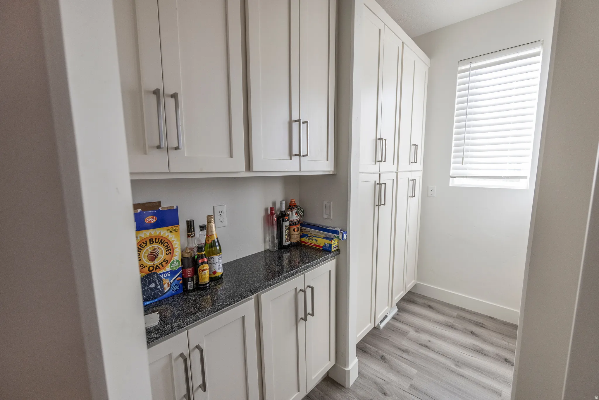 White shaker-style cabinetry with brushed nickel hardware, expansive built-in pantry, dark flecked countertop, and wood-finish flooring