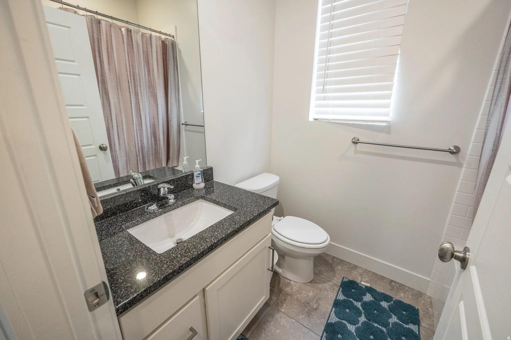 White vanity with dark speckled countertop and integrated sink, chrome faucet, tiled flooring, and a window with blinds