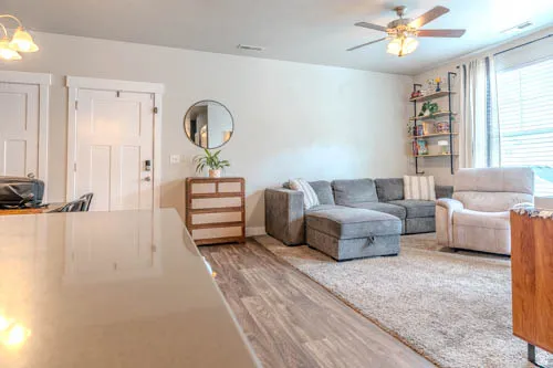 Open-concept living space featuring wood-finish flooring throughout, a gray upholstered sectional with storage ottoman, ceiling fan with integrated lighting, round mirror accent, wooden dresser, wall-mounted shelving, and abundant natural light from multi