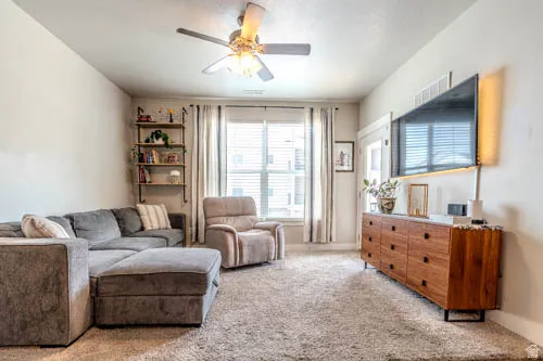 Bright and spacious living room featuring neutral wall finishes, ceiling fan with integrated lighting, large windows with dual curtain panels, engineered wood-finish flooring, and neutral area rug