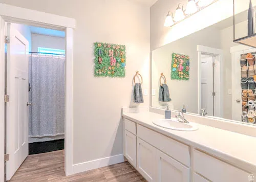 Spa-inspired primary bathroom featuring dual vanities with ample countertop space, large framing mirrors with integrated lighting, wood-finish flooring, and access to an ensuite shower area