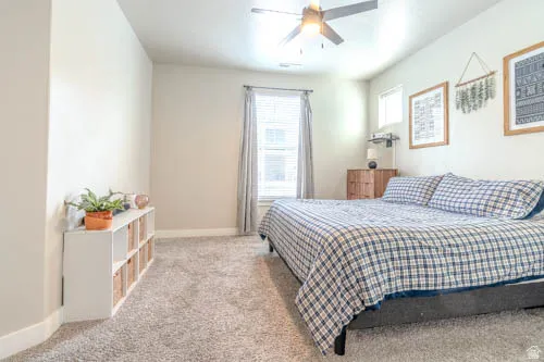 Spacious primary bedroom with plush carpeting, abundant natural light from dual windows with soft furnishings, ceiling fan with integrated lighting, and ample floor space
