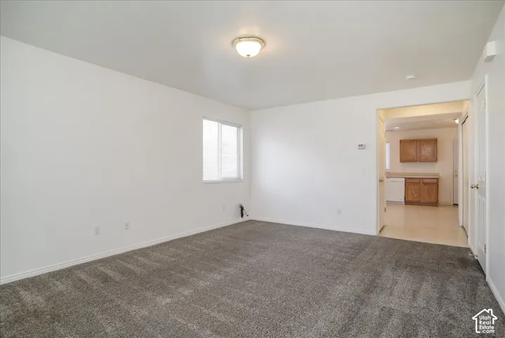 Carpeted flooring, white walls, ceiling-mounted light fixture, window with blinds, and an entryway to a kitchen area with wood cabinets and light-colored countertops