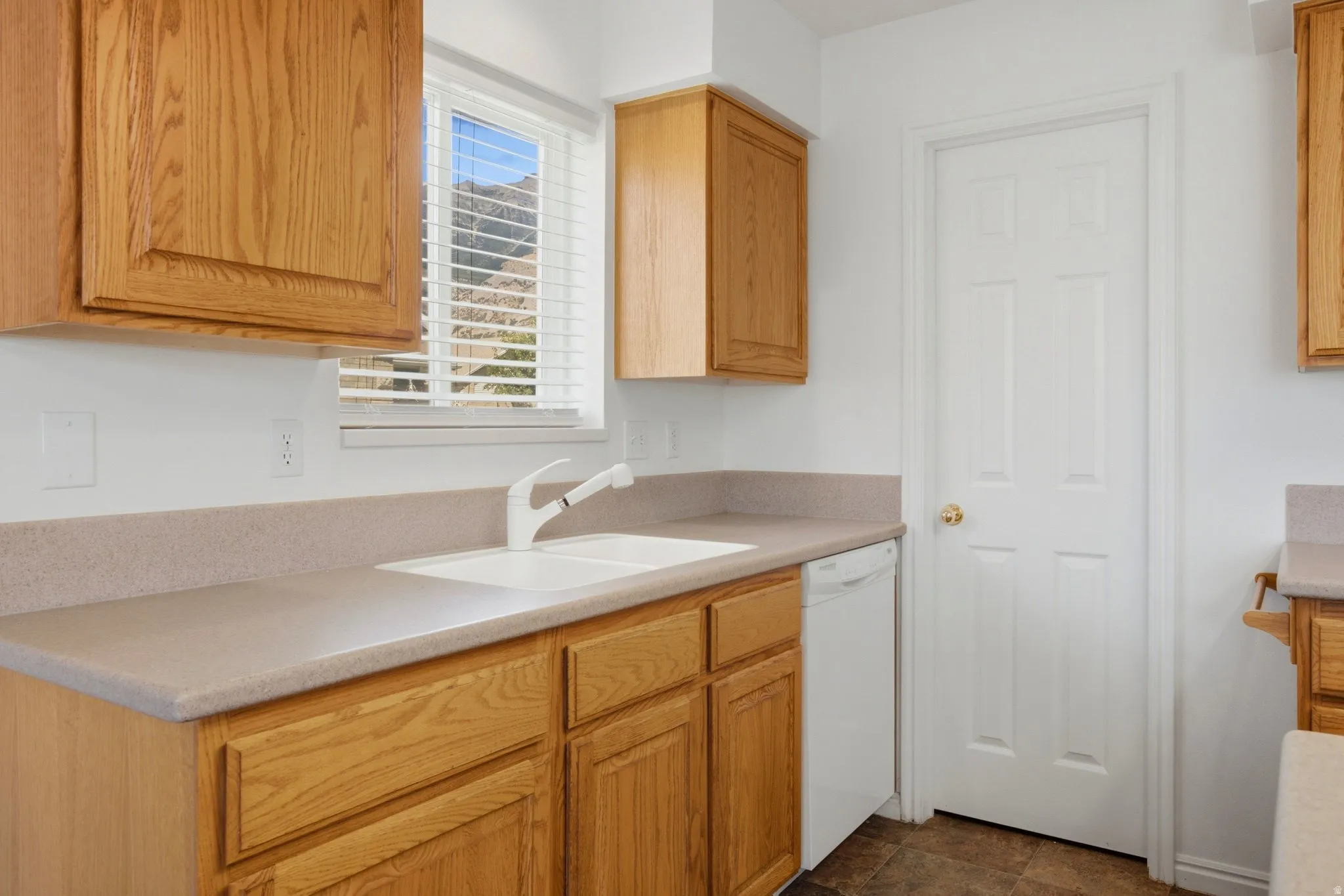 Wood cabinetry with a double basin sink, white dishwasher, and light-colored countertops