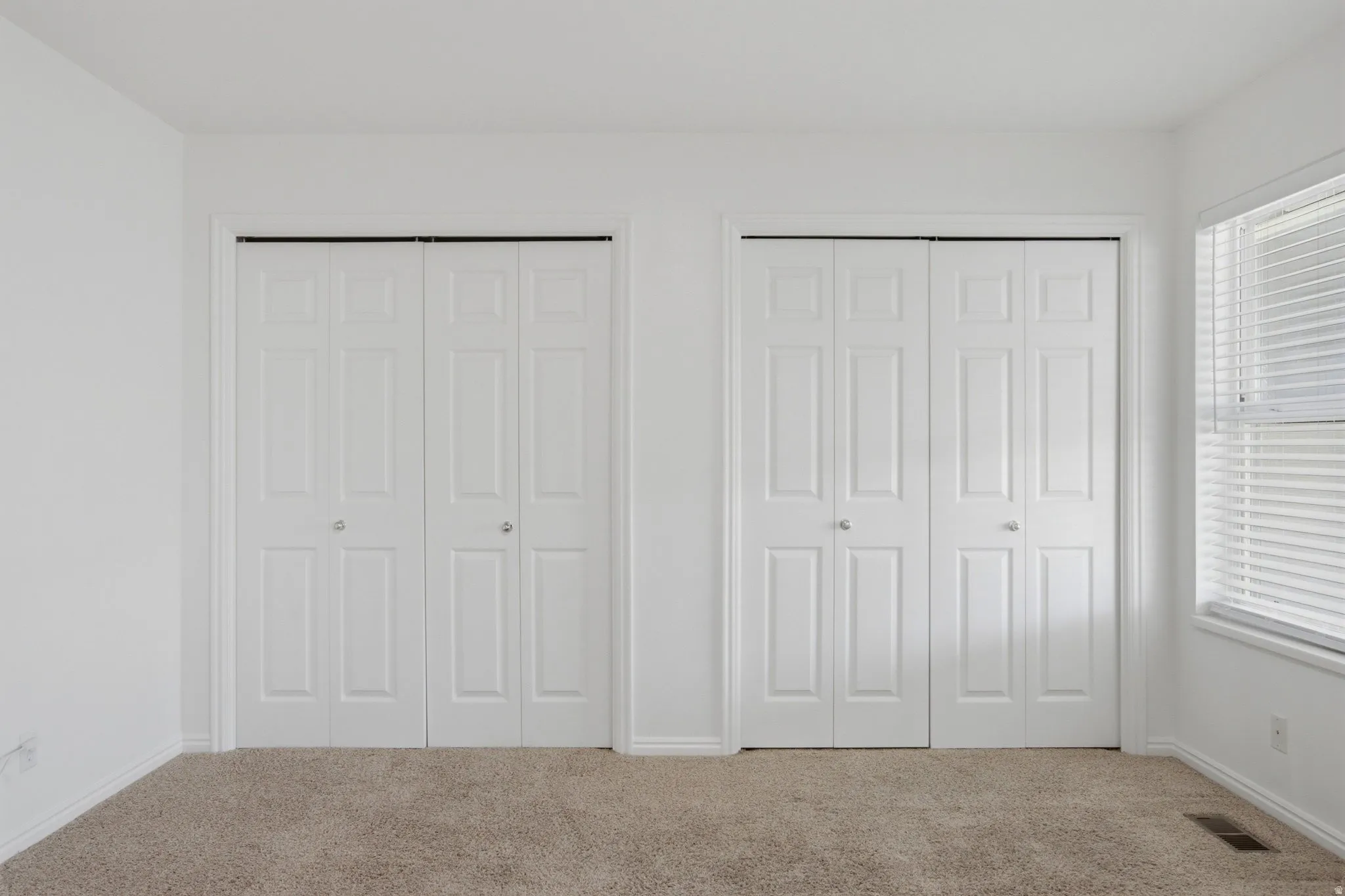 Dual bi-fold closet doors, neutral wall paint, window with blinds, and carpeting