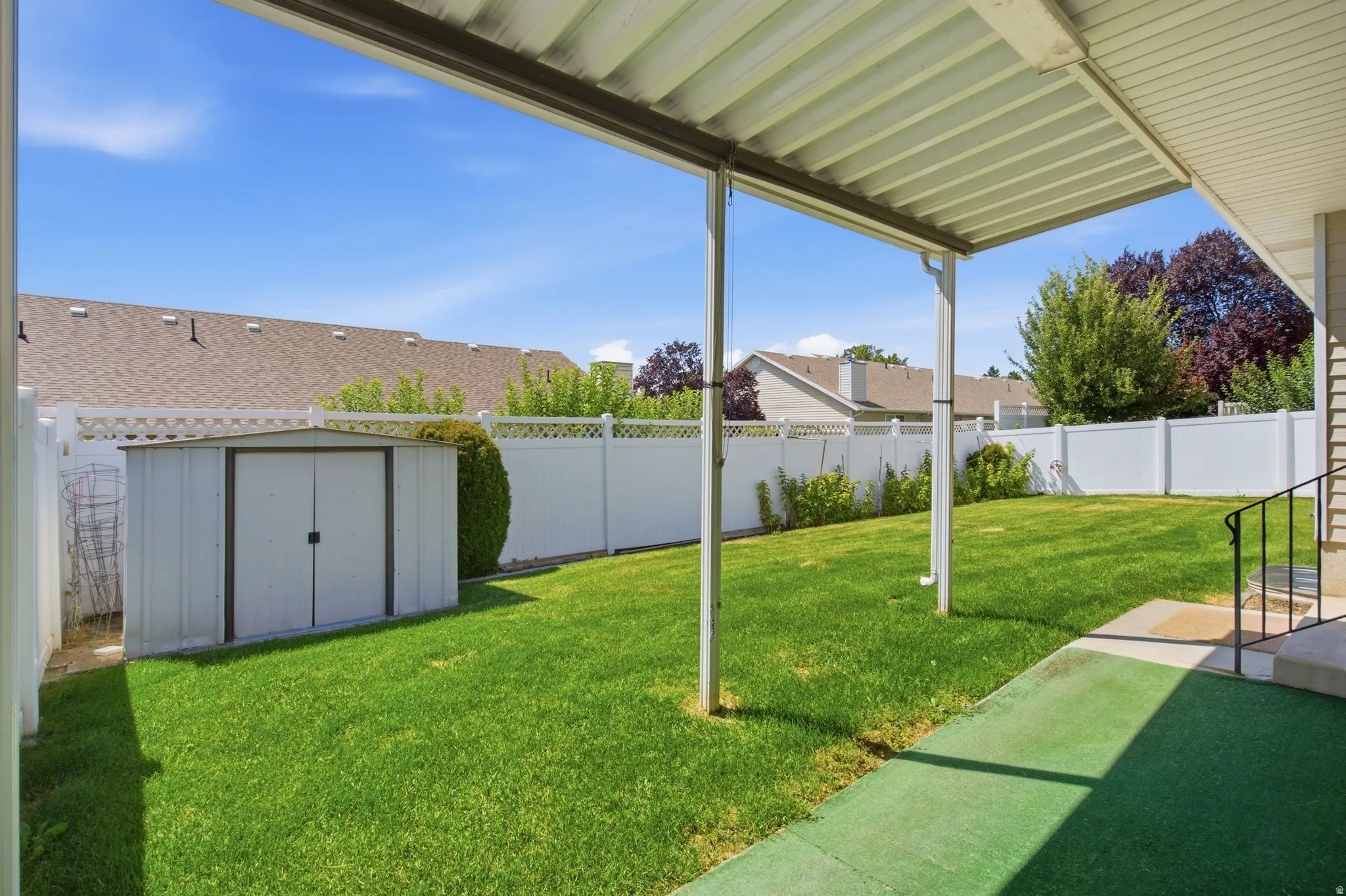 Covered patio with an artificial turf surface, a spacious lawn, a white privacy fence, and an exterior storage shed