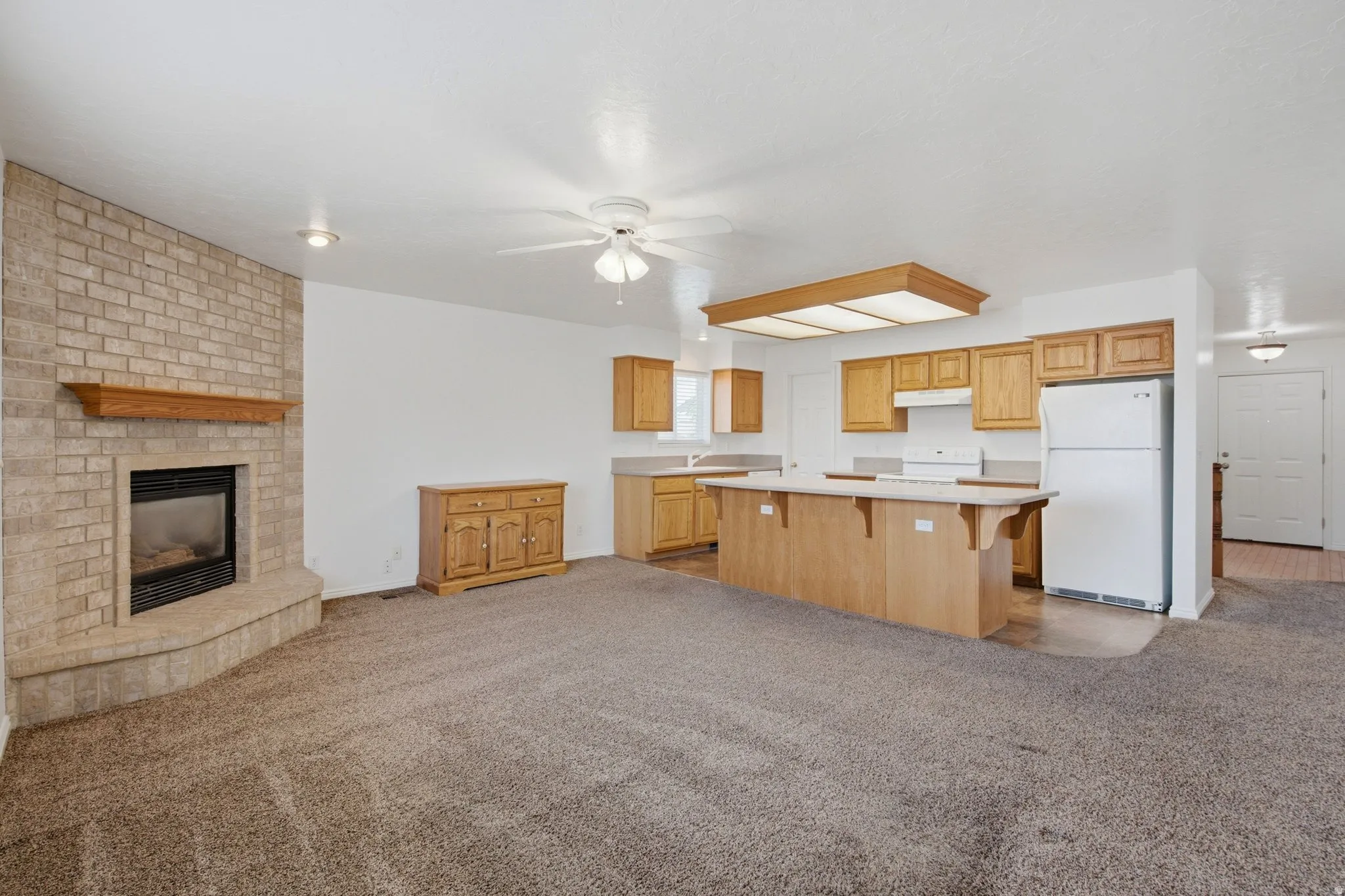 Brick-faced fireplace with a wood mantel, wood-finish kitchen cabinetry, kitchen island with counter seating, ceiling fan, and recessed lighting