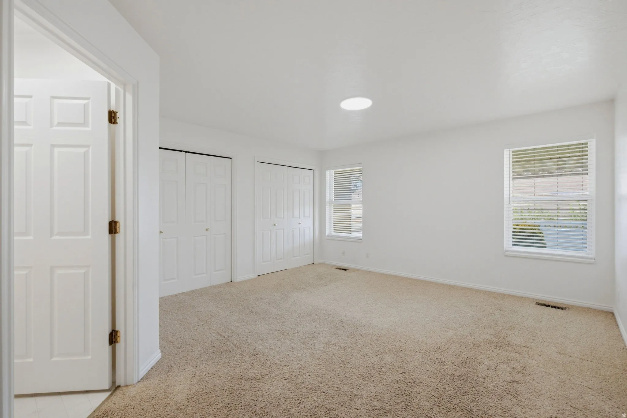 Neutral carpet flooring, white walls, two bi-fold closet doors, and two windows with horizontal blinds