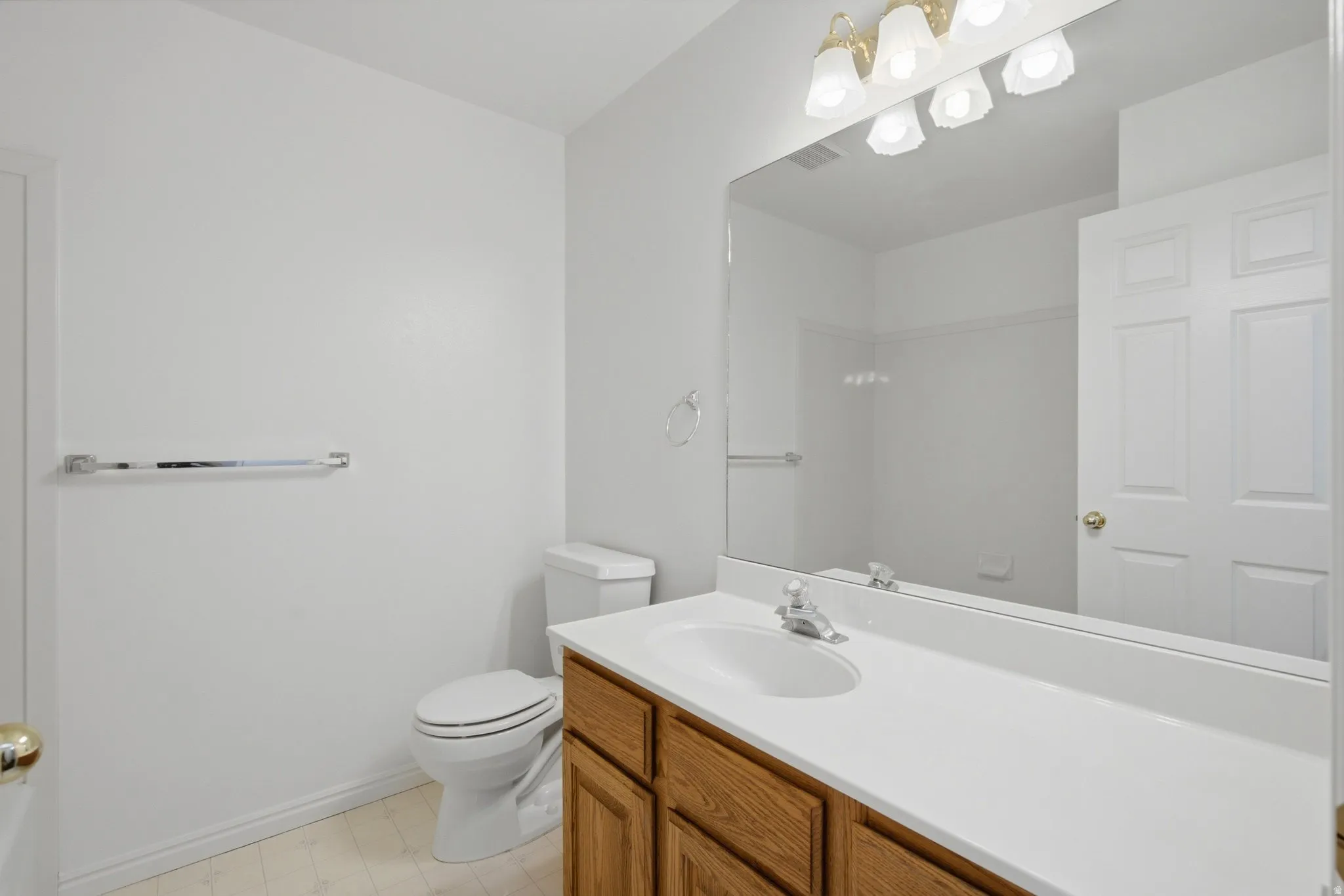 Vanity with wood-finish cabinetry and integrated sink, large wall-mounted mirror, overhead vanity lighting, and white toilet