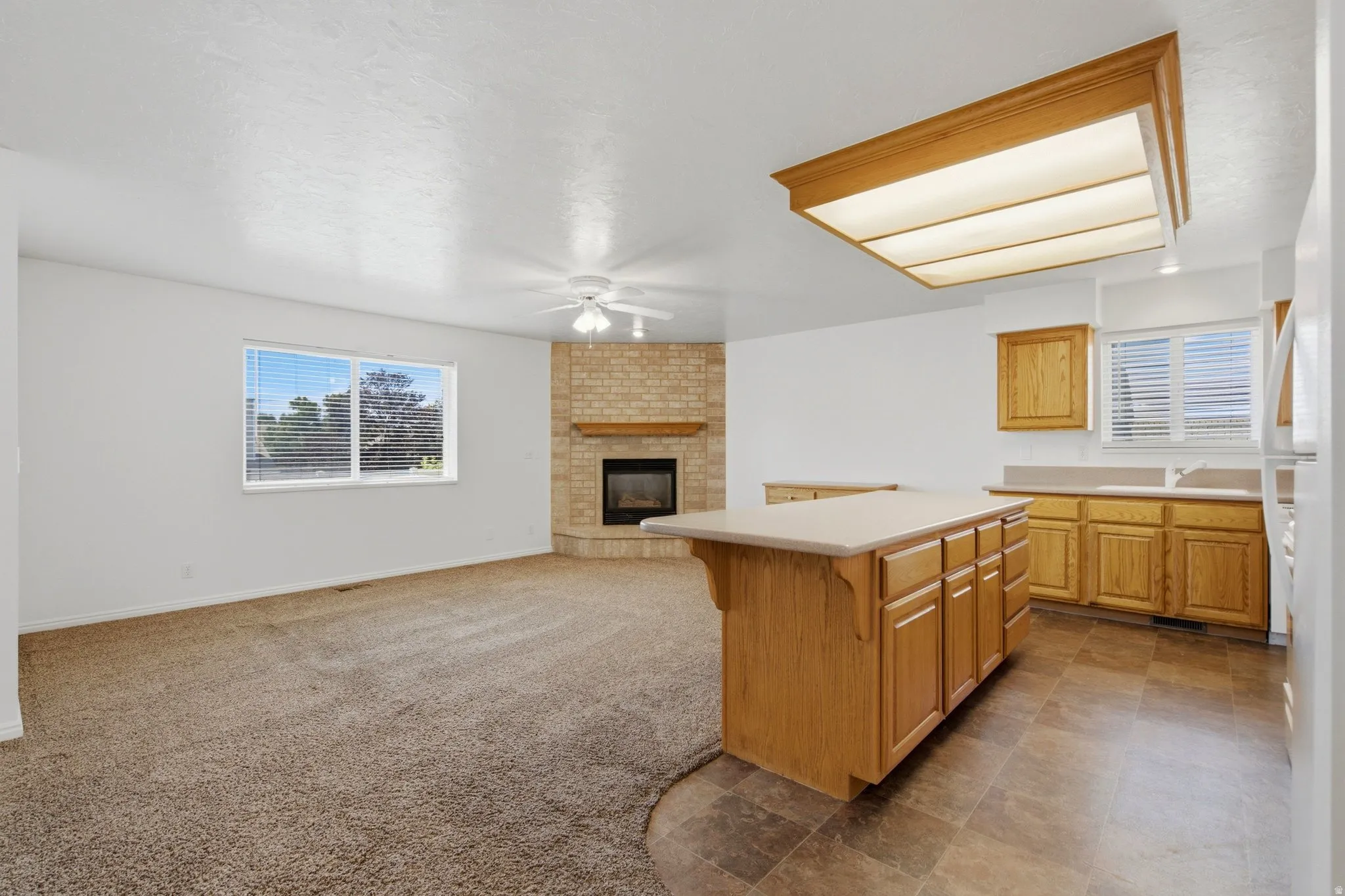 Open-concept living space featuring a corner fireplace with brick surround, ceiling fan, and window with horizontal blinds