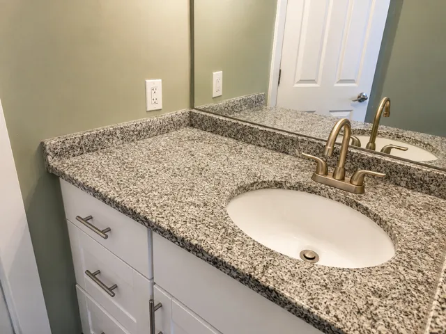 White vanity with shaker-style cabinet doors, brushed nickel hardware, a speckled countertop, and an undermount sink with a brass-finish faucet