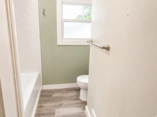White bathtub with textured tile surround, wood-finish flooring, white window trim, and a wall-mounted towel bar