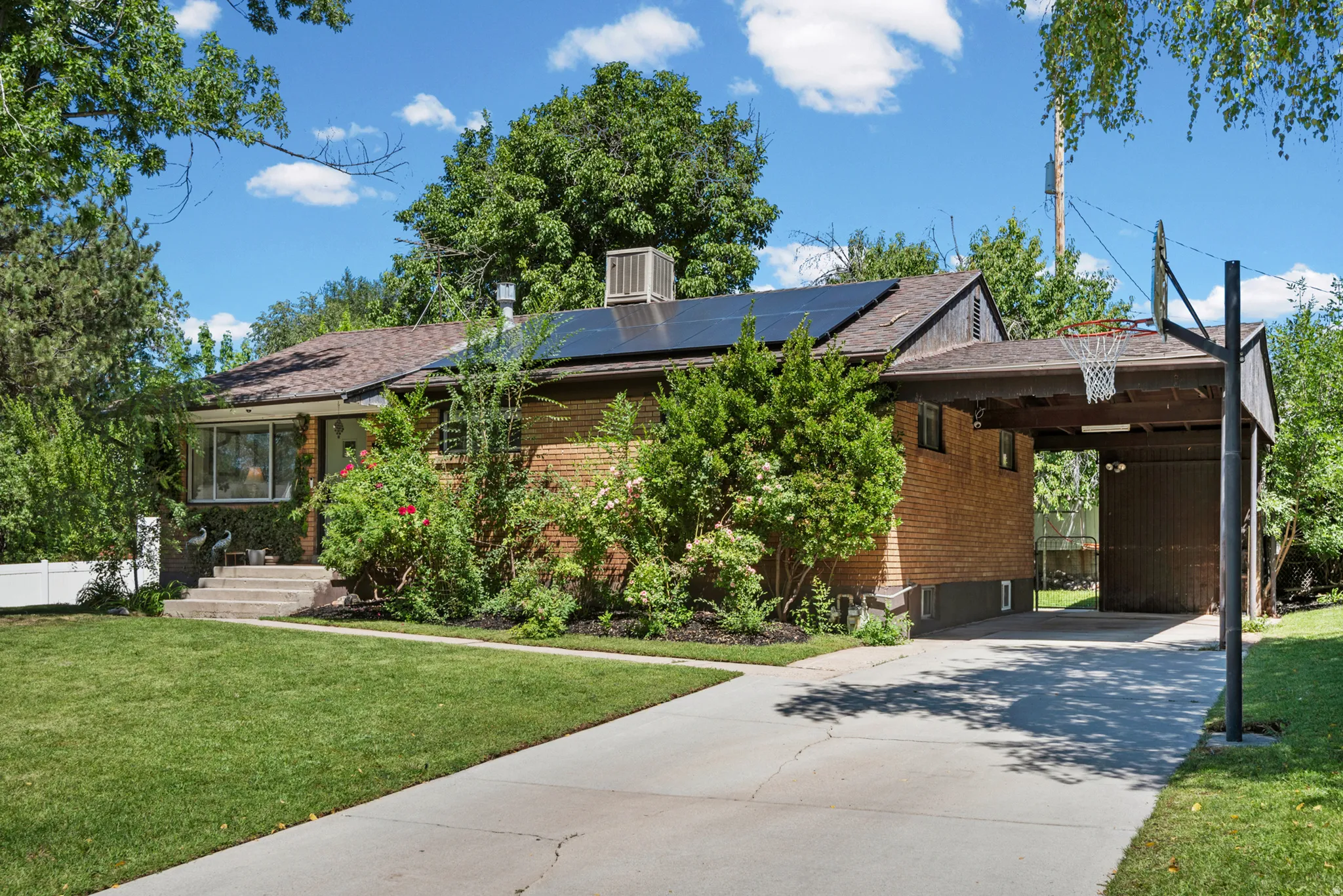 Brick exterior with solar panels, covered carport, concrete driveway, and mature landscaping